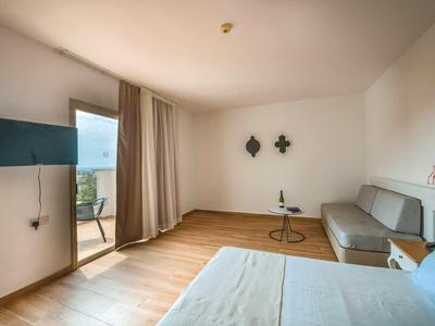 Helles Hotelzimmer mit Bett, grauer Couch, kleinem Tisch, TV an der Wand und Balkon mit Ausblick.