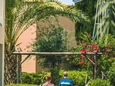 Zwei Personen gehen auf einem Gartenweg entlang von grünen Pflanzen und Palmen.