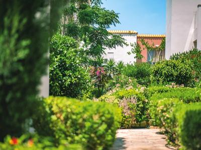 Weg mit grünen Sträuchern und Bäumen entlang einer weißen Gebäudefassade bei sonnigem Wetter.