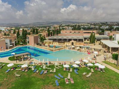 Großer Außenpool mit Liegestühlen auf grünem Rasen vor Gebäuden unter bewölktem Himmel.
