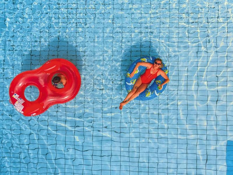 Zwei Personen schwimmen im blauen Pool, eine auf einem roten Schwimmring, die andere schwimmt frei.