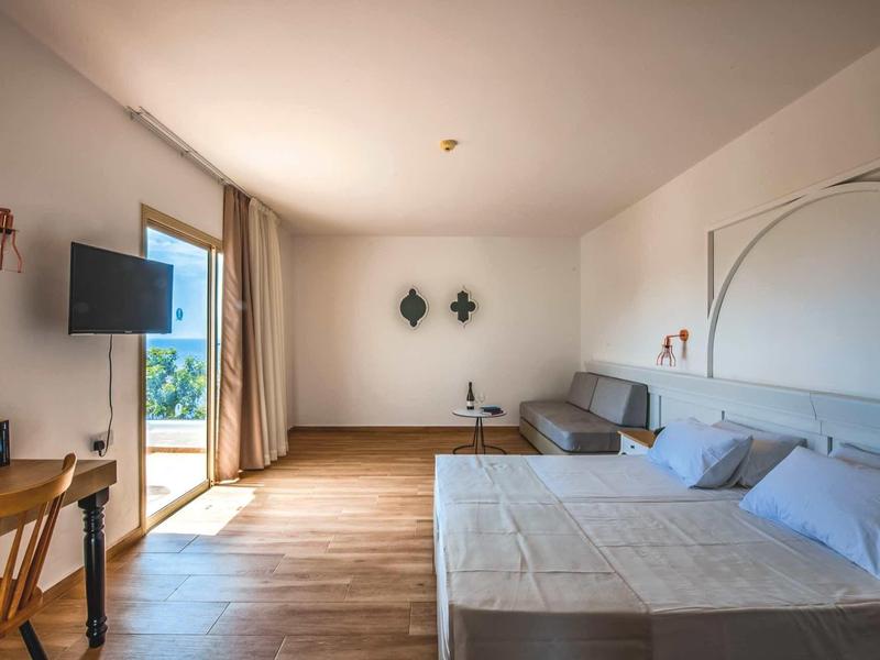 Helles, modernes Hotelzimmer mit Doppelbett, Sofa, Schreibtisch, TV und Balkon mit Meeresblick.