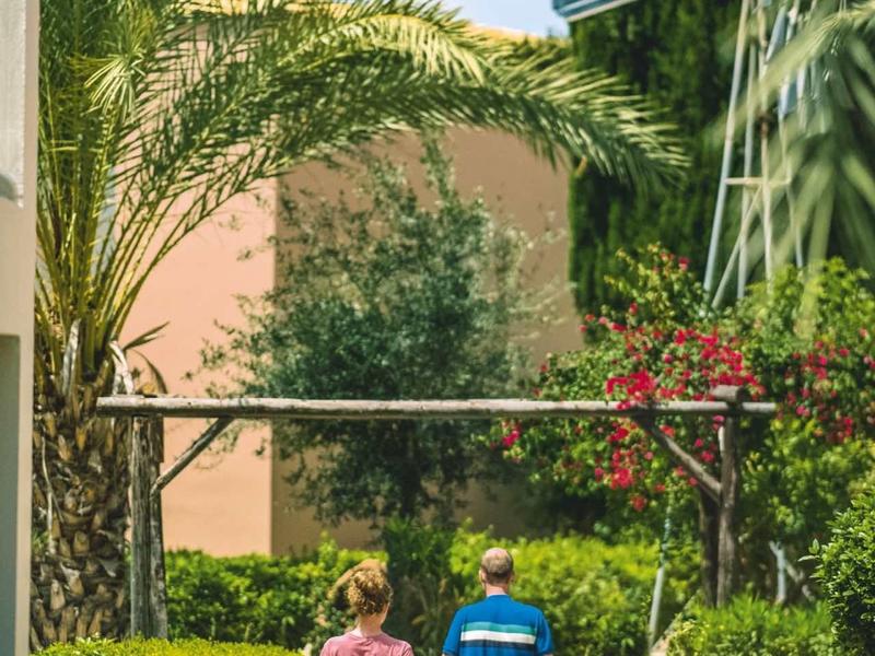 Zwei Kinder gehen auf einem schmalen Gartenweg, umgeben von grünen Pflanzen und einer Palme.