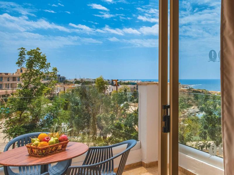 Balkon mit Tisch, Stühlen und Obstkorb, Blick auf grüne Bäume und blauen Himmel am Meer.