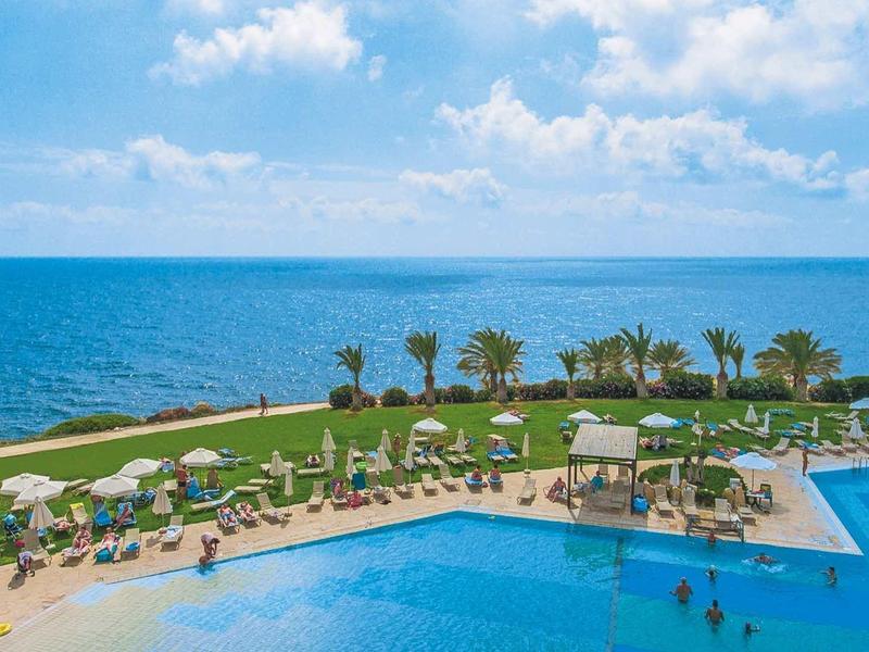 Großer blauer Pool mit Sonnenliegen, Palmen und Blick auf ruhiges Meer unter blauem Himmel.