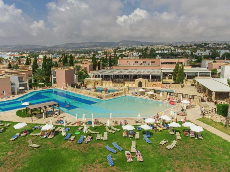 Großer Außenpool mit Liegestühlen auf grünem Rasen vor Gebäuden unter bewölktem Himmel.