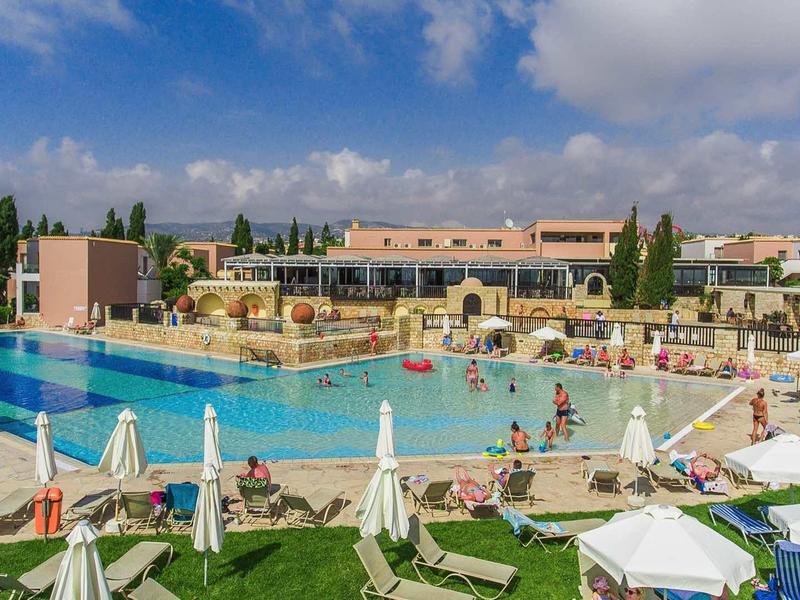 Großer Außenpool mit Sonnenschirmen, Liegen und Menschen, umgeben von Hotelgebäuden und grünen Bäumen.