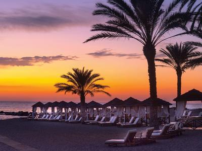 Sonnenuntergang am Strand mit Liegestühlen, Sonnenschirmen und Palmen.