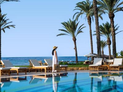 Poolbereich mit Liegestühlen und Palmen neben dem Meer an einem sonnigen Tag.