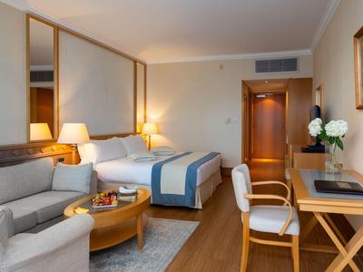 Modern hotel room with double bed, sofa, desk, and wooden flooring.