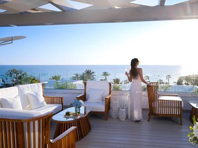 A woman enjoys the sea view from a terrace with wooden furniture at a hotel.