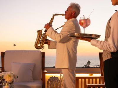 A saxophonist in a white suit plays on a terrace at sunset with a sea view.