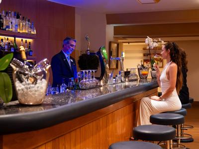Woman in white dress sitting at hotel bar, bartender serving drinks, warm lighting