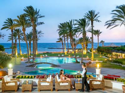 Luxurious hotel with illuminated pool, palm trees, and sea view at sunset.