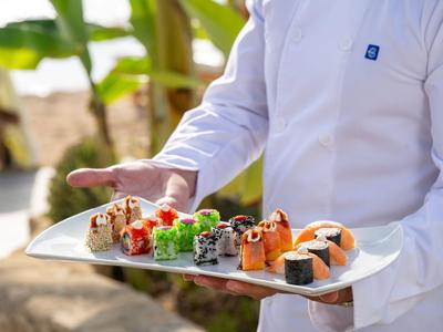 A waiter holds a tray with various sushi types in a sunny outdoor setting.