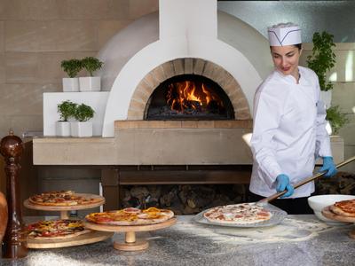 A chef prepares pizza in a hotel kitchen in front of a wood-fired oven with flames.