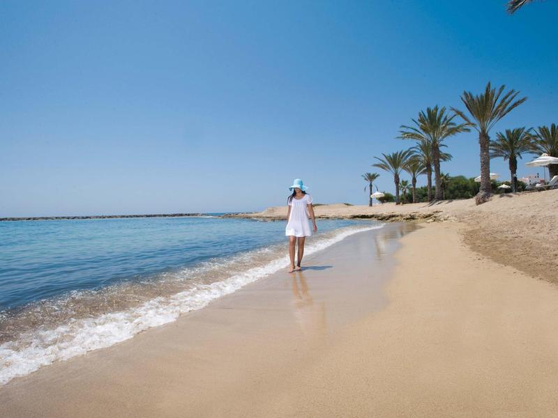 Frau im weißen Kleid geht am Sandstrand entlang, blauer Himmel und Palmen im Hintergrund.