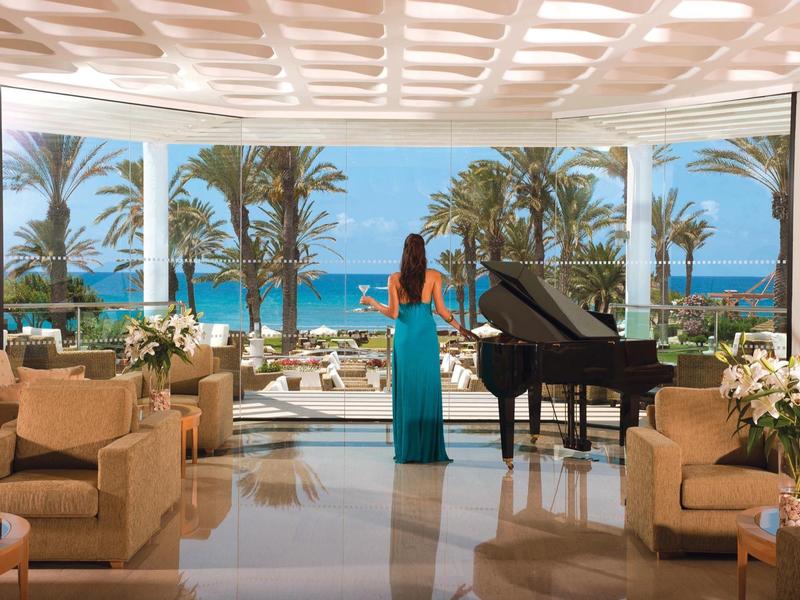 Eleganter Raum mit Klavier, zwei Sofas und großer Glasfront mit Meerblick und Palmen im Hintergrund.