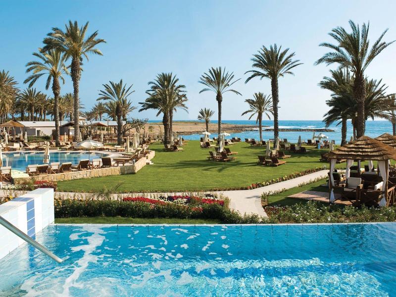 Ein tropischer Poolbereich mit Palmen, Liegestühlen und Blick auf das blaue Meer im Hintergrund.