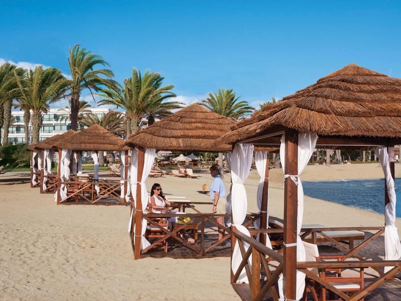 Strand mit Strohdach-Pavillons, Sand, Palmen und blauem Himmel im Hintergrund.