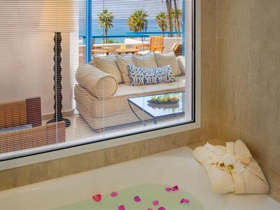 Innenansicht eines Hotels mit Badewanne, die mit Blütenblättern gefüllt ist, und Blick auf eine Terrasse mit Meerblick.