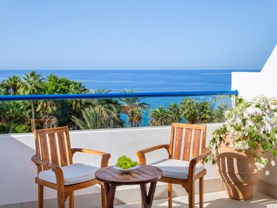 Balcone con due sedie in legno, tavolo e vista sul mare e palme.