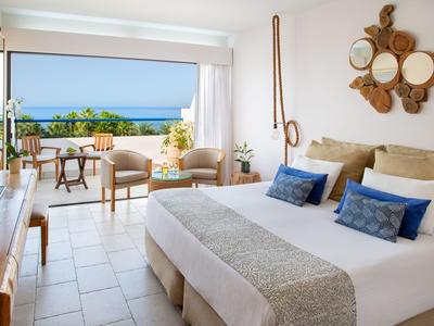 Una camera d'hotel luminosa con letto matrimoniale, zona salotto e balcone con vista sul mare.