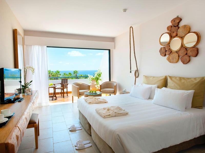 Hell eingerichtetes Hotelzimmer mit zwei Betten, Balkon und Meerblick, sanfte Naturtöne dominieren.