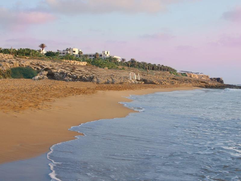 Ein ruhiger Strand mit goldenem Sand, felsiger Küste und blauem Wasser unter leicht bewölktem Himmel.