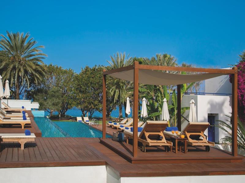 Luxuriöser Poolbereich mit Sonnenliegen, Holzsteg und Palmen unter klarem blauem Himmel.