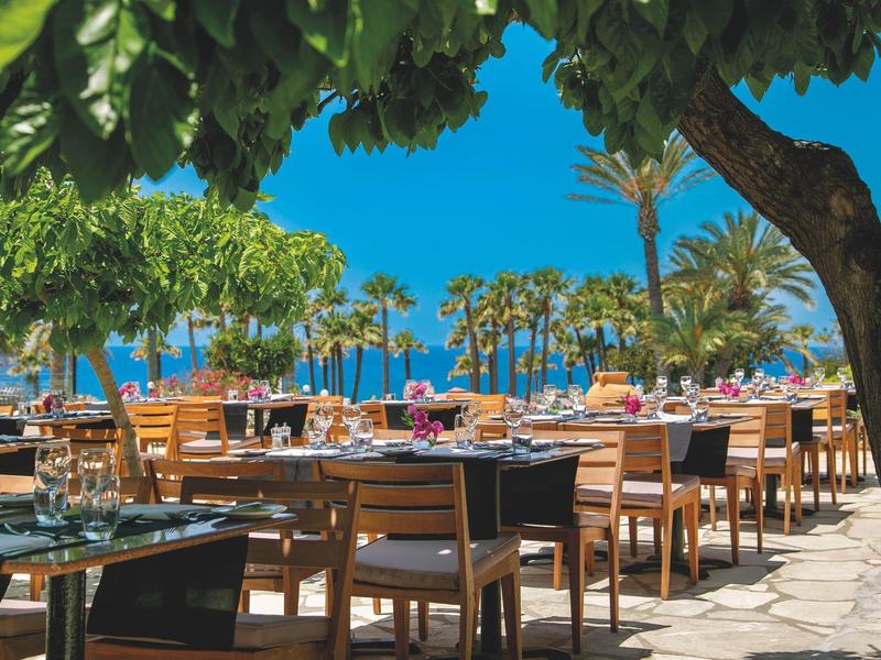 Außenbereich eines Restaurants mit Holztischen und Stühlen, umgeben von Palmen und blauem Himmel.