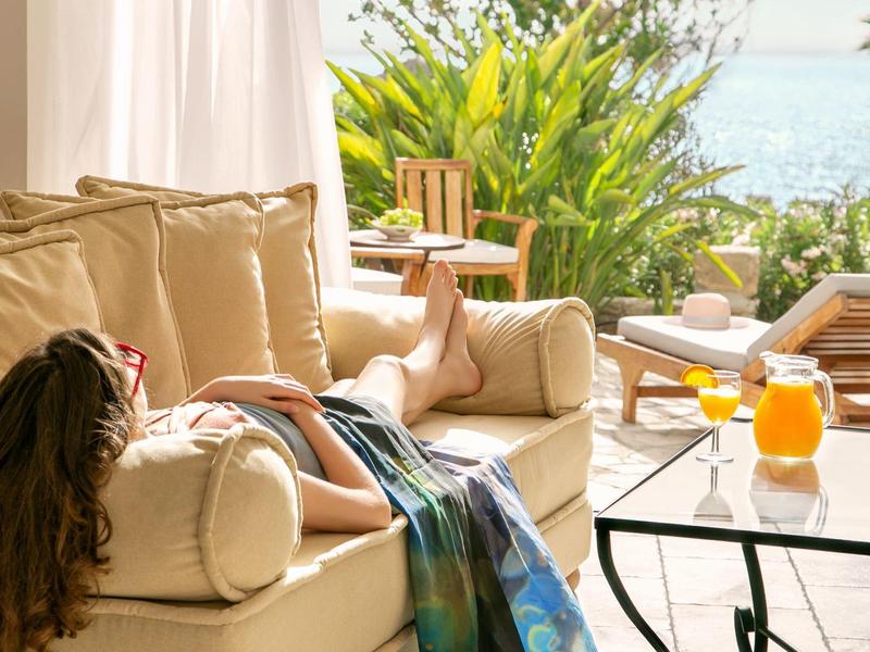 Frau entspannt auf hellem Sofa im hellen Raum mit Blick auf Garten und Meer, Glas Orangensaft auf Tisch.