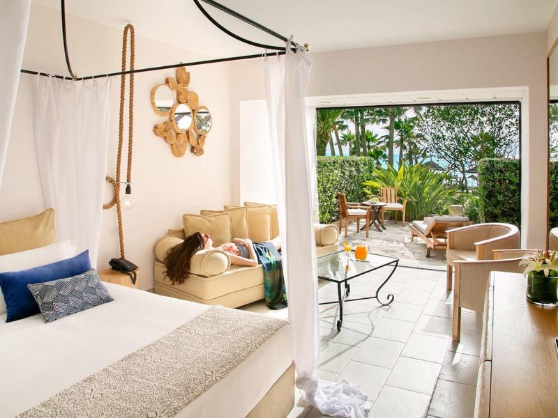 Helles Schlafzimmer mit weißem Bett, gemütlicher Sitzecke und offener Tür zu Balkon mit Grünblick.