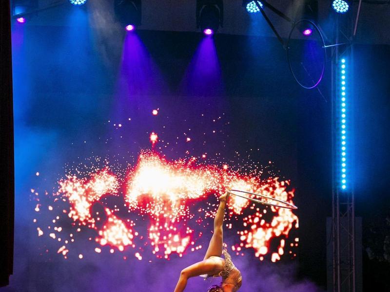 Tänzer macht Handstand auf Bühne mit buntem Bühnenlicht und Feuer-Effekten im Hintergrund.