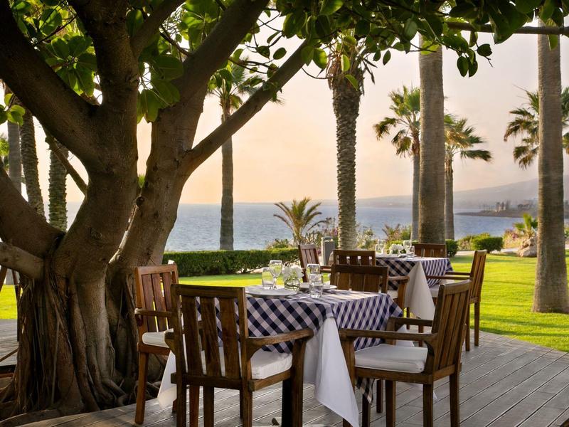 Terrasse mit Tischen und Stühlen, umgeben von Palmen und Blick auf das Meer bei Sonnenuntergang.