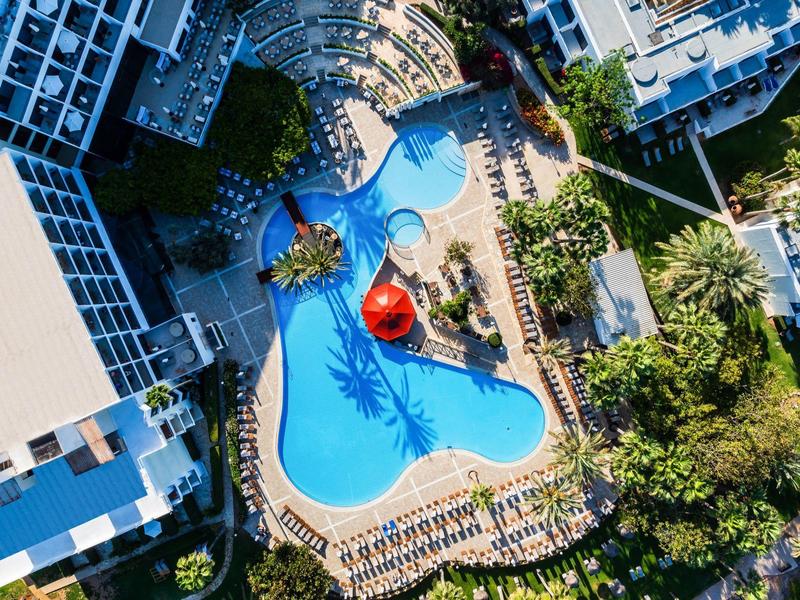 Luftaufnahme eines Hotels mit großem, organisch geformtem blauen Pool und Sonnenliegen.