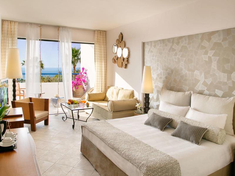 Lichte hotelkamer met groot bed, zithoek en balkon met uitzicht op zee.