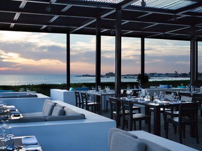 Offenes Restaurant mit weißen Sitzsofas und Tischen mit Blick auf den Sonnenuntergang über dem Meer.