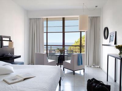 Habitación de hotel luminosa con cama, sillas y vista al mar a través de grandes ventanas.