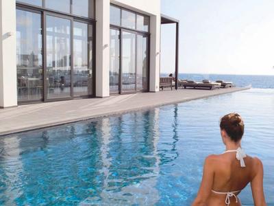 Mujer en bikini blanco entrando a una piscina con vista al mar junto a un edificio moderno.