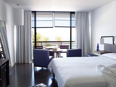 Habitación de hotel luminosa con cama doble, dos sillones y balcón con vista al exterior.