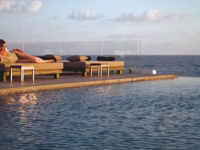 Persona descansando en una tumbona junto a una piscina infinita con vista al mar al atardecer.