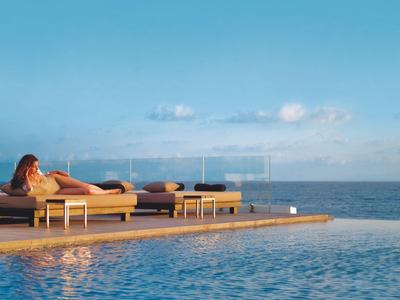 Frau entspannt auf Sonnenliege am Poolrand mit Blick auf ruhiges Meer und blauem Himmel.