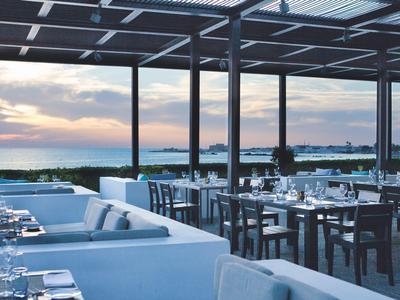 Offenes Restaurant mit weißen Sofas, Holztischen und Blick auf Sonnenuntergang über dem Meer.