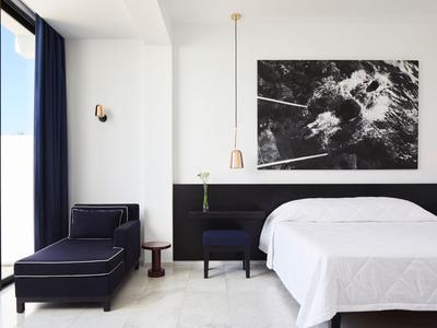 Helles Hotelzimmer mit großem Bett, dunklem Sessel, Kunst an der Wand und Glasbalkontür.