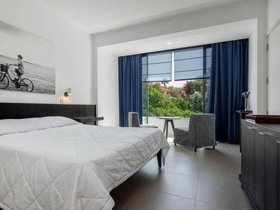 Helles Schlafzimmer mit Doppelbett, weißen Wänden, blauen Vorhängen und Blick auf Garten.