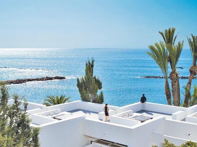 Moderne weiße Gebäude mit Terrasse, Palmen und Meer im Hintergrund unter klarem Himmel.