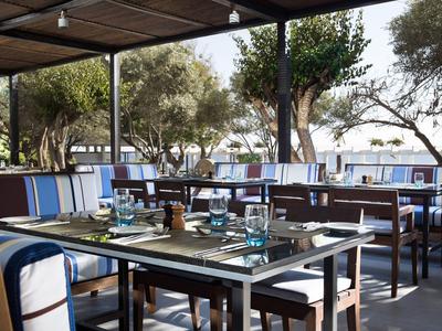 Offenes Restaurant mit Holztischen, blauen Sitzpolstern und Blick auf Bäume und Meer.