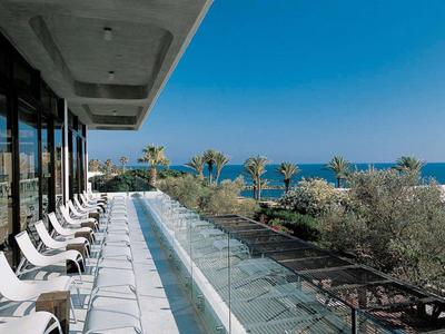 Balkon mit weißen Stühlen, Glasgeländer, Blick auf Palmen und Meer unter blauem Himmel.