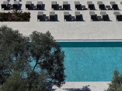 Luftaufnahme eines blauen Hotelpools mit schwarzen Liegestühlen und grünen Bäumen im Vordergrund.